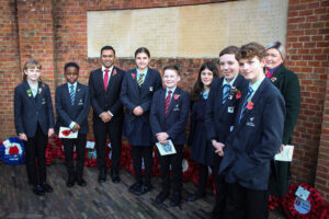 A group photo of students and adults, all wearing poppies, standing in front of a brick wall memorial inscribed with names, surrounded by poppy wreaths at the base.