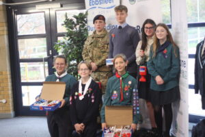 A group of young people, including a soldier or cadet, students, and members of a youth organization, pose indoors. Several are wearing poppy emblems and holding boxes of poppies for a Poppy Appeal fundraiser.