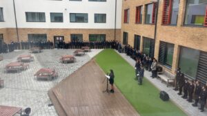 A group of six students in school uniform and two adults pose for a photo outdoors on a small turf stage. Everyone is wearing a poppy emblem, and one student holds a trumpet.