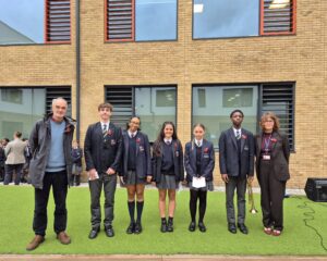 A group of six students in school uniform and two adults pose for a photo outdoors on a small turf stage. Everyone is wearing a poppy emblem, and one student holds a trumpet.