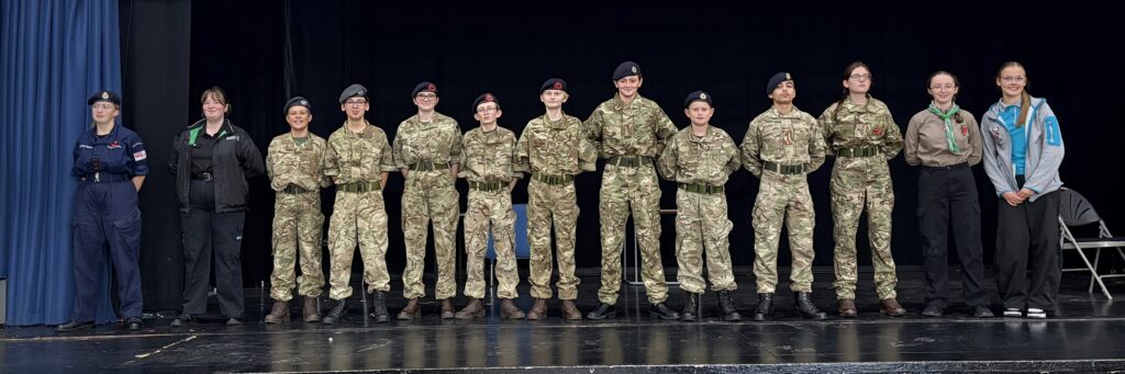 A line of eleven young people standing on a stage against a black backdrop, wearing various uniforms including military camouflage, cadet, and youth organization uniforms.