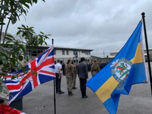 Outdoor parade ground with people in military and formal attire standing in formation, framed by a Union Jack flag and a blue and yellow crested flag.