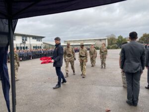 An outdoor military ceremony. A man in a coat walks to the left, carrying a poppy wreath, followed by a line of cadets in camouflage uniform, while other students stand in formation in the background.