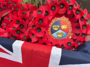 A close-up of a large poppy wreath with a central circular crest for the Combined Cadet Force, resting on a folded Union Jack flag.