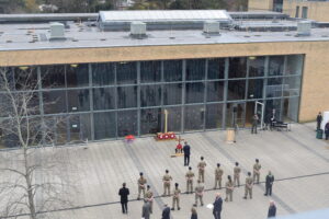 A high-angle wide shot of a large outdoor Remembrance ceremony. A formation of cadets and officials stand on the plaza, facing a display of a wooden cross and wreaths, while a large crowd watches from inside the glass-fronted building.