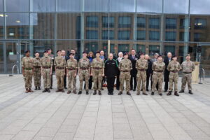 A formal group photo taken outdoors with a modern building reflected in the glass background. The group is composed of a line of people in military camouflage and cadet uniforms, mixed with several adults in formal attire.