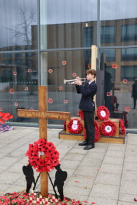A student in school uniform plays a trumpet next to a wooden commemorative cross and a display of poppy wreaths and soldier silhouettes outside a modern building.