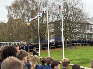 An outdoor Remembrance ceremony at a school. A woman speaks at a microphone in the foreground, facing rows of students in uniform who stand by a line of flags bearing military silhouettes.
