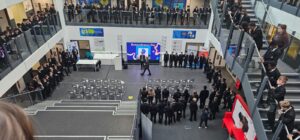 A wide-angle shot of a Remembrance Day assembly held in a large, multi-story school atrium. Students line the stairs and balconies, watching a presentation on a screen below.