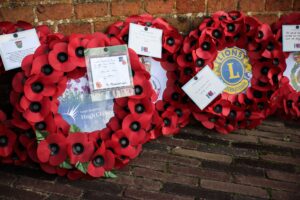 Poppy wreaths for Remembrance Day laid against a brick wall. The wreaths, featuring bright red poppies, include tributes from Leigh Academy and Lions Clubs International.