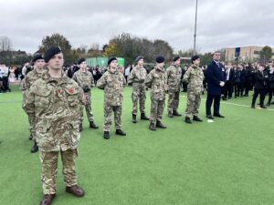 A line of young cadets in camouflage uniforms and a man in a dark coat stand at attention on a green sports field during an outdoor ceremony, with a crowd of students in the background.