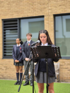 A young woman in a school uniform, with a poppy pin, speaks into a microphone from a music stand on an outdoor stage. Two other students stand in the background.